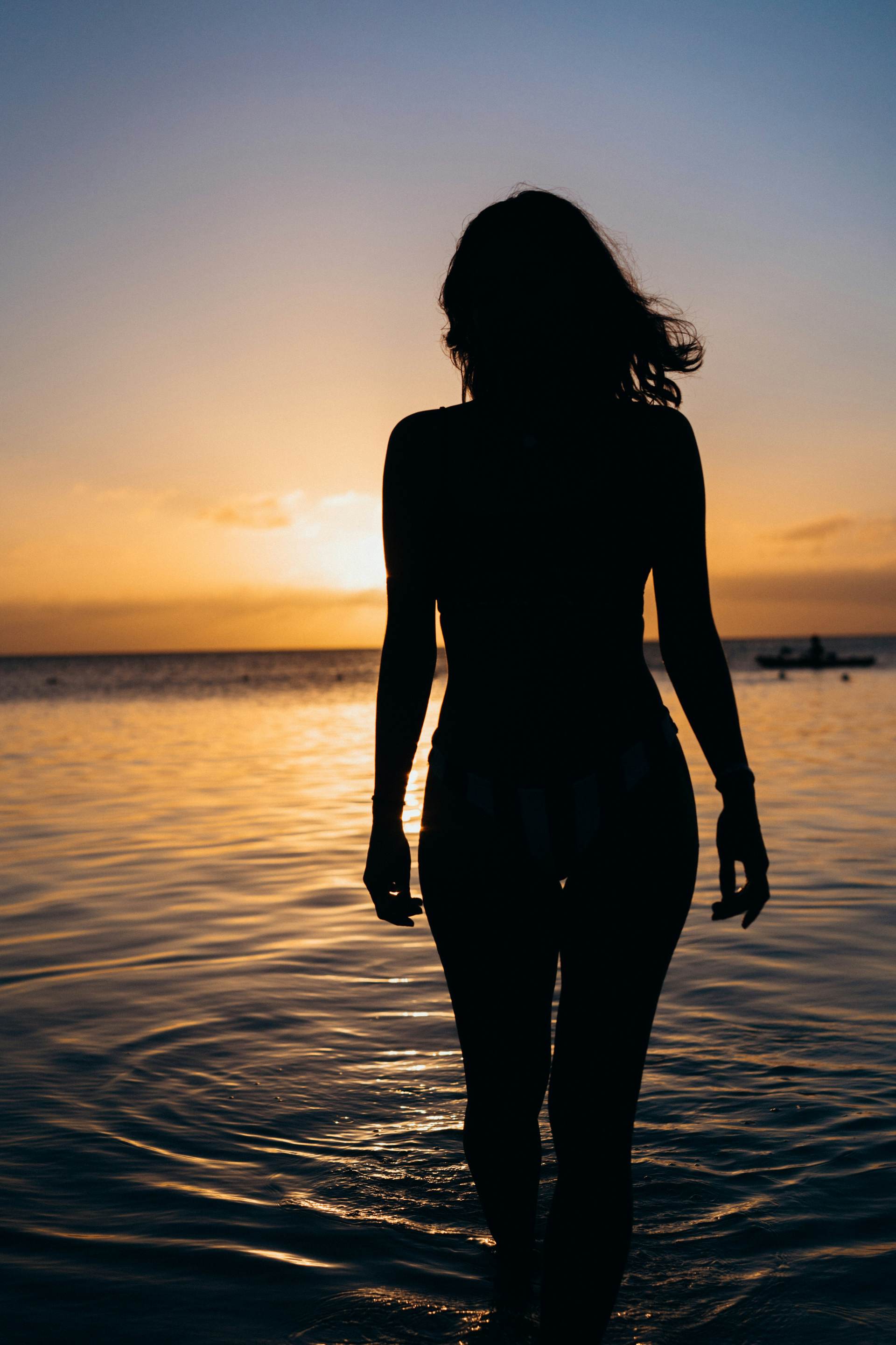 A woman silhouetted at the edge of a golden ocean at dusk, facing the water
